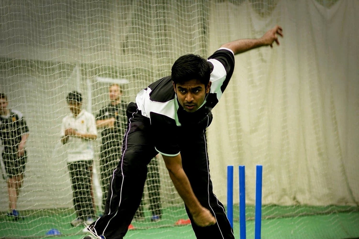 A young bowler in an indoor net, with juniors and a coach watching from behind the netting